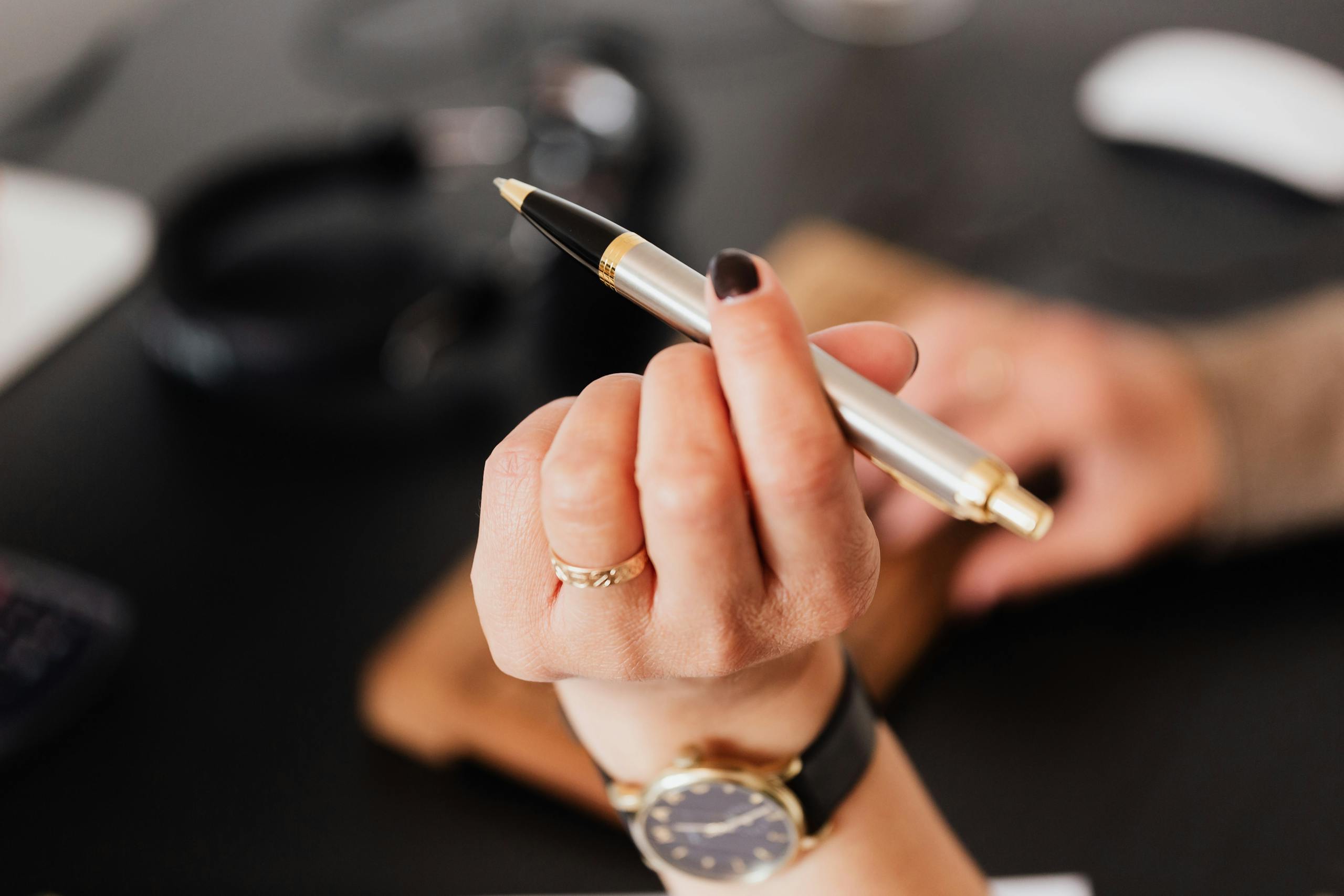 A close-up of a hand holding a silver pen, set against a blurred office background.