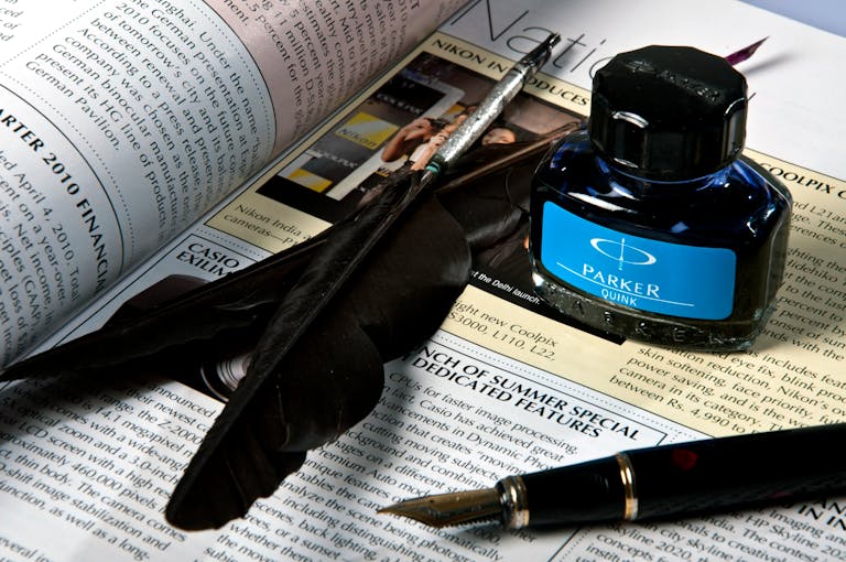 Close-up of feather, ink bottle, and pen resting on an opened newspaper page.