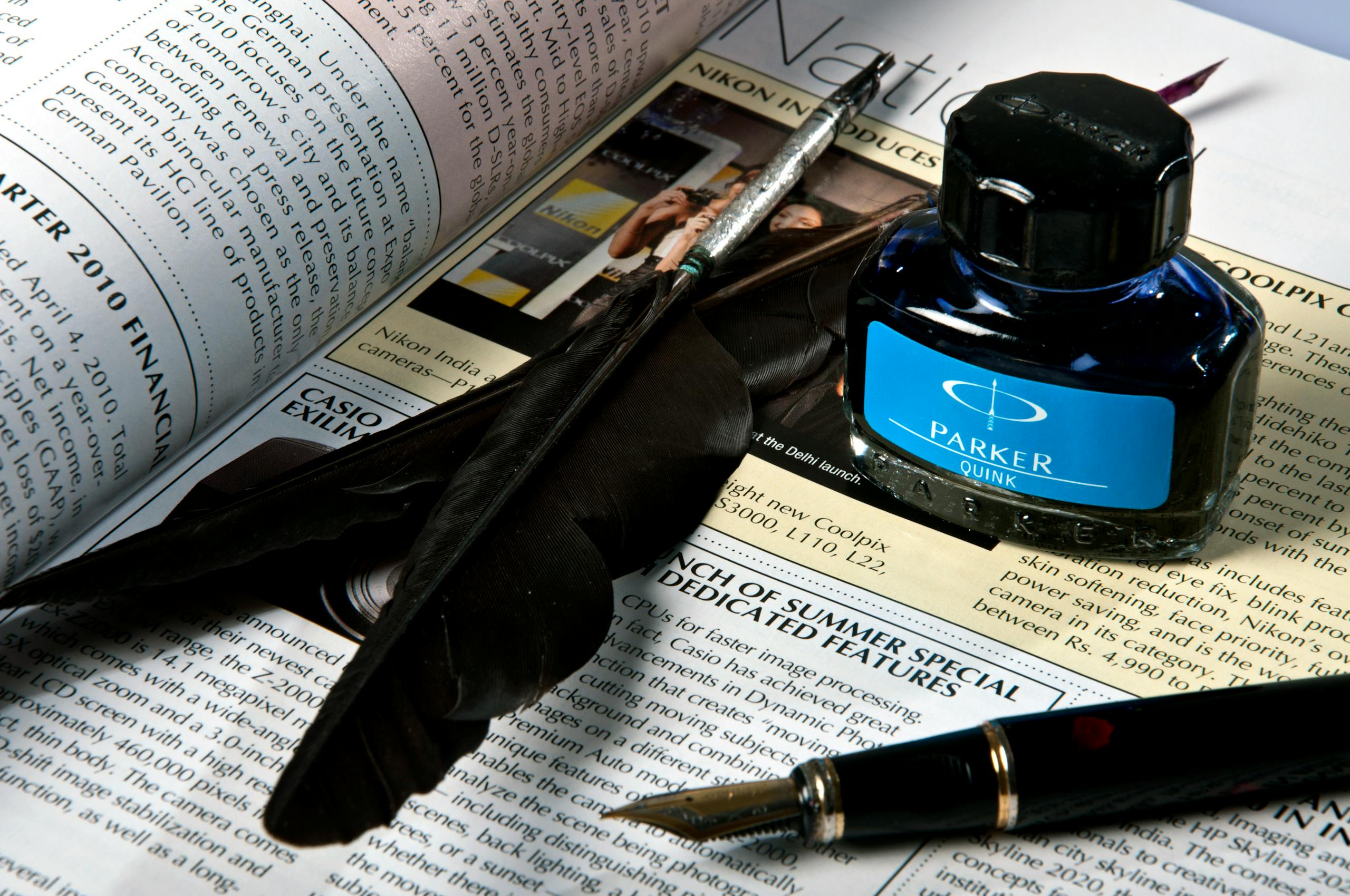 Close-up of feather, ink bottle, and pen resting on an opened newspaper page.