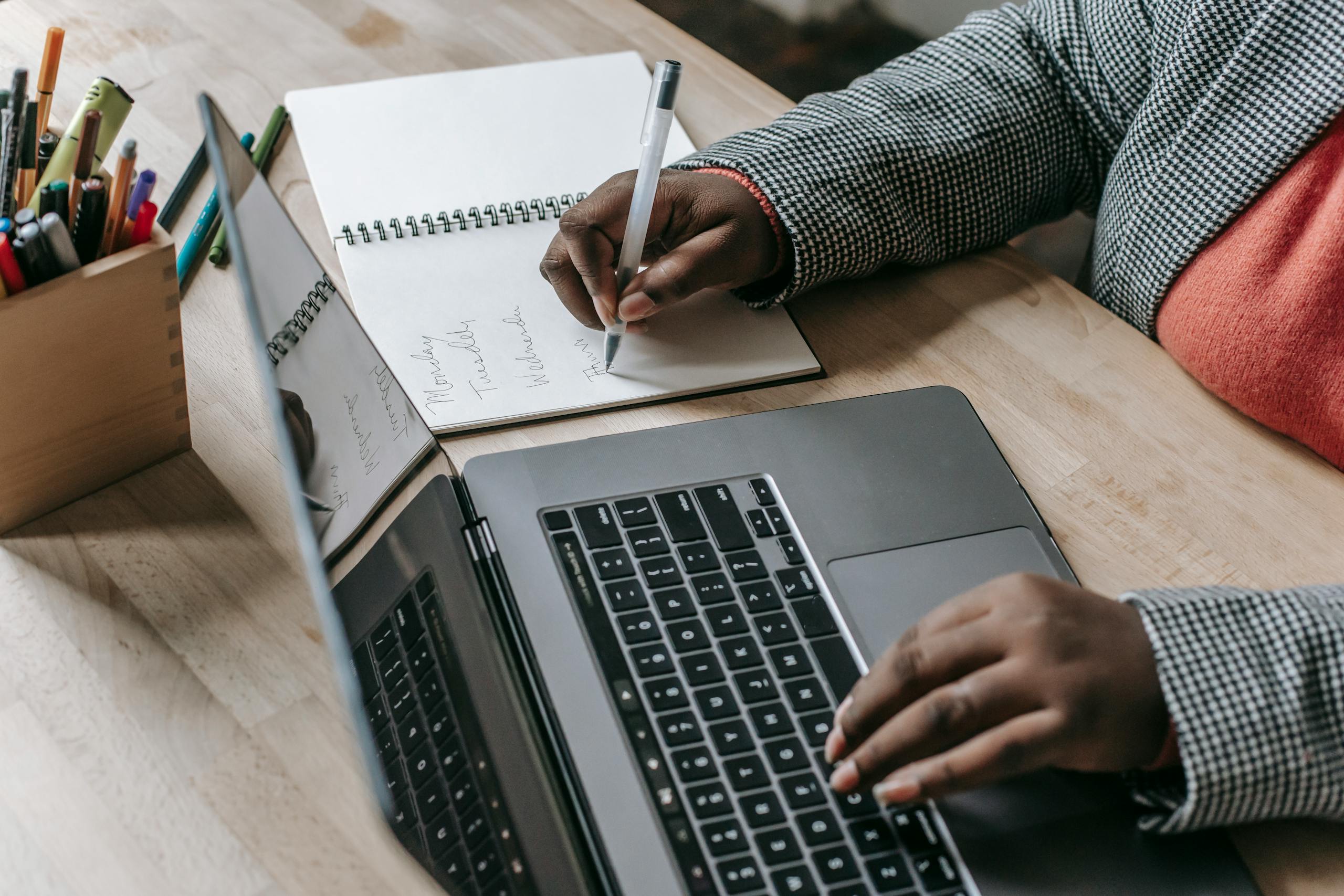 Crop unrecognizable African American female in formal wear writing notes in notepad while working on laptop